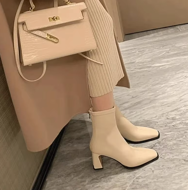 A pair of beige, square-toed ankle boots with high heels, worn by a person with a matching handbag.
